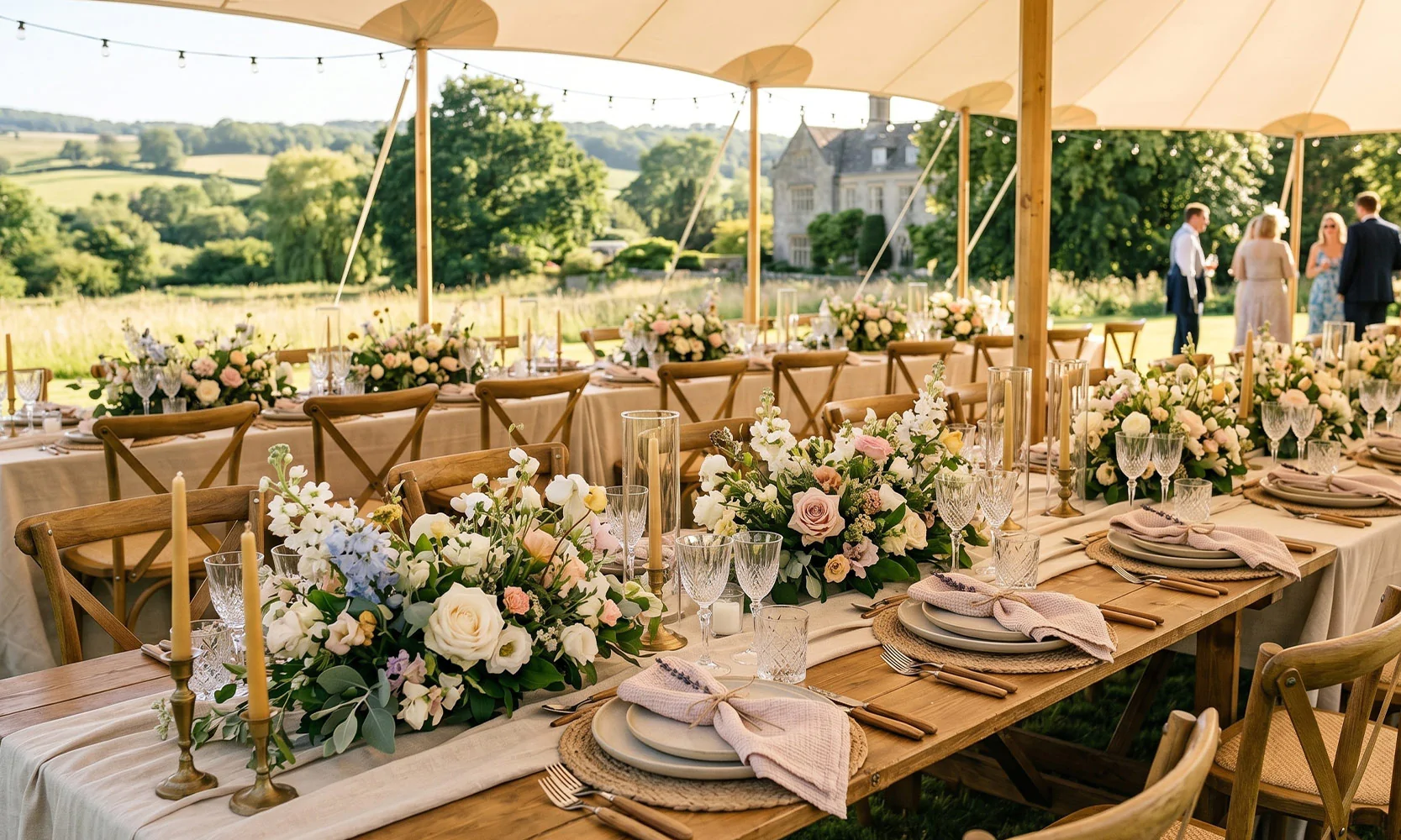 Elegant outdoor wedding dining setup under a sailcloth marquee featuring long wooden banquet tables, neutral linen runners, layered place settings with pink napkins, and lush seasonal floral centerpieces.