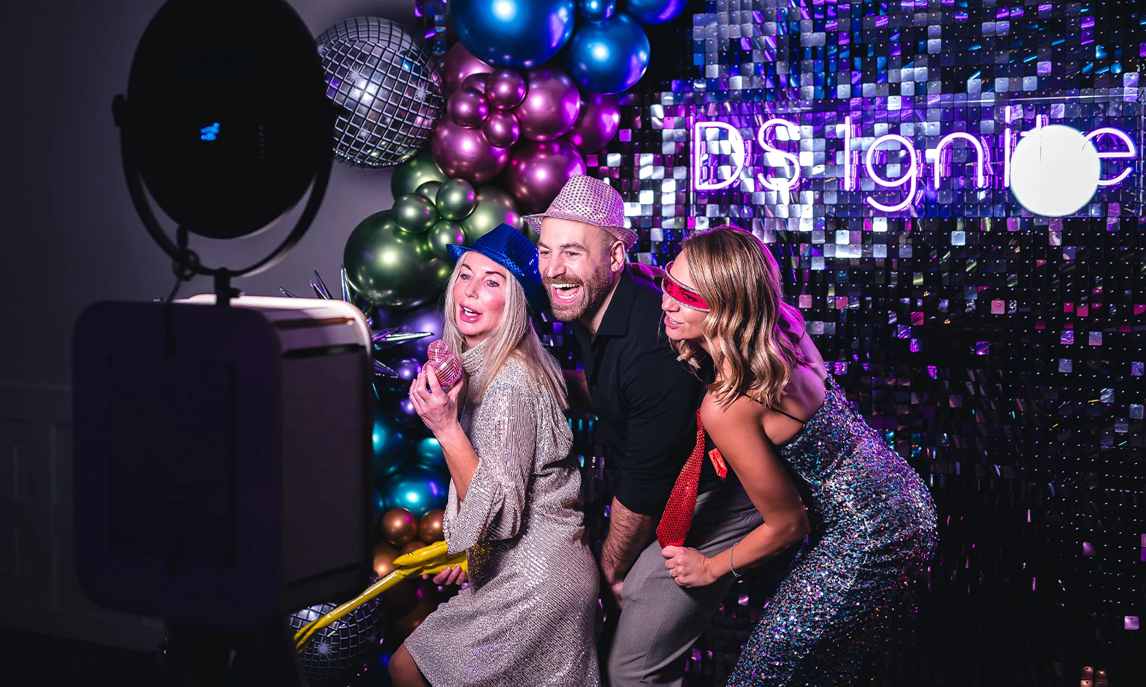Guests posing at a corporate event with an unmanned photo booth with a sequin wall and custom neon sign backdrop.