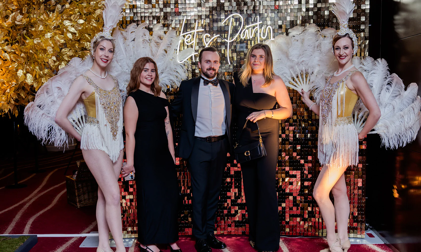 Guests posing for photos with burlesque dancers at a corporate event with silver sequin wall and neon sign.